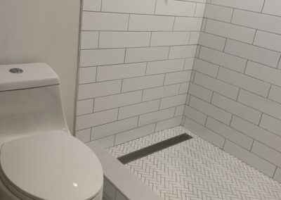 A modern bathroom corner with a white ceramic toilet next to a walk-in shower area with white subway tiles and herringbone-patterned floor tiles.