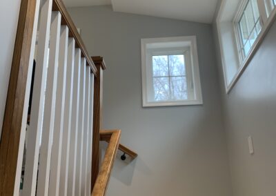 A staircase with wooden handrails and white balusters leading to a hallway with gray walls and a window providing natural light.