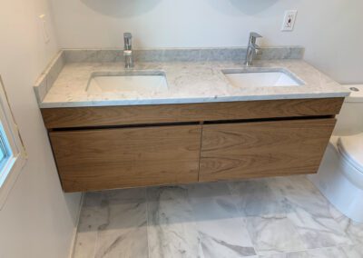 A modern bathroom vanity with a marble countertop featuring two sinks, set against a white wall with a toilet to the right.