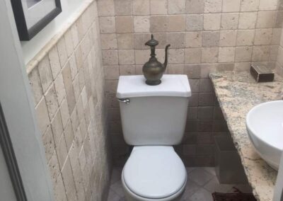 A small bathroom featuring a toilet with a decorative piece on the tank, tiled walls, and artwork hanging above.