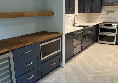 A modern kitchen featuring navy blue cabinets, wooden countertops, stainless steel appliances, open wooden shelving, and herringbone patterned tile flooring.